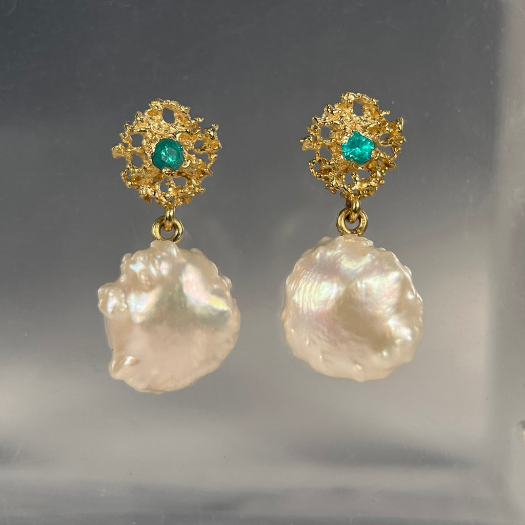photo shows a pair of pearl earrings; the pearls are roundish but bumpy and irregular; they drop from a lacy, textured gold piece that is also roundish but smaller in diameter than the pearls and has a single emerald in the center 