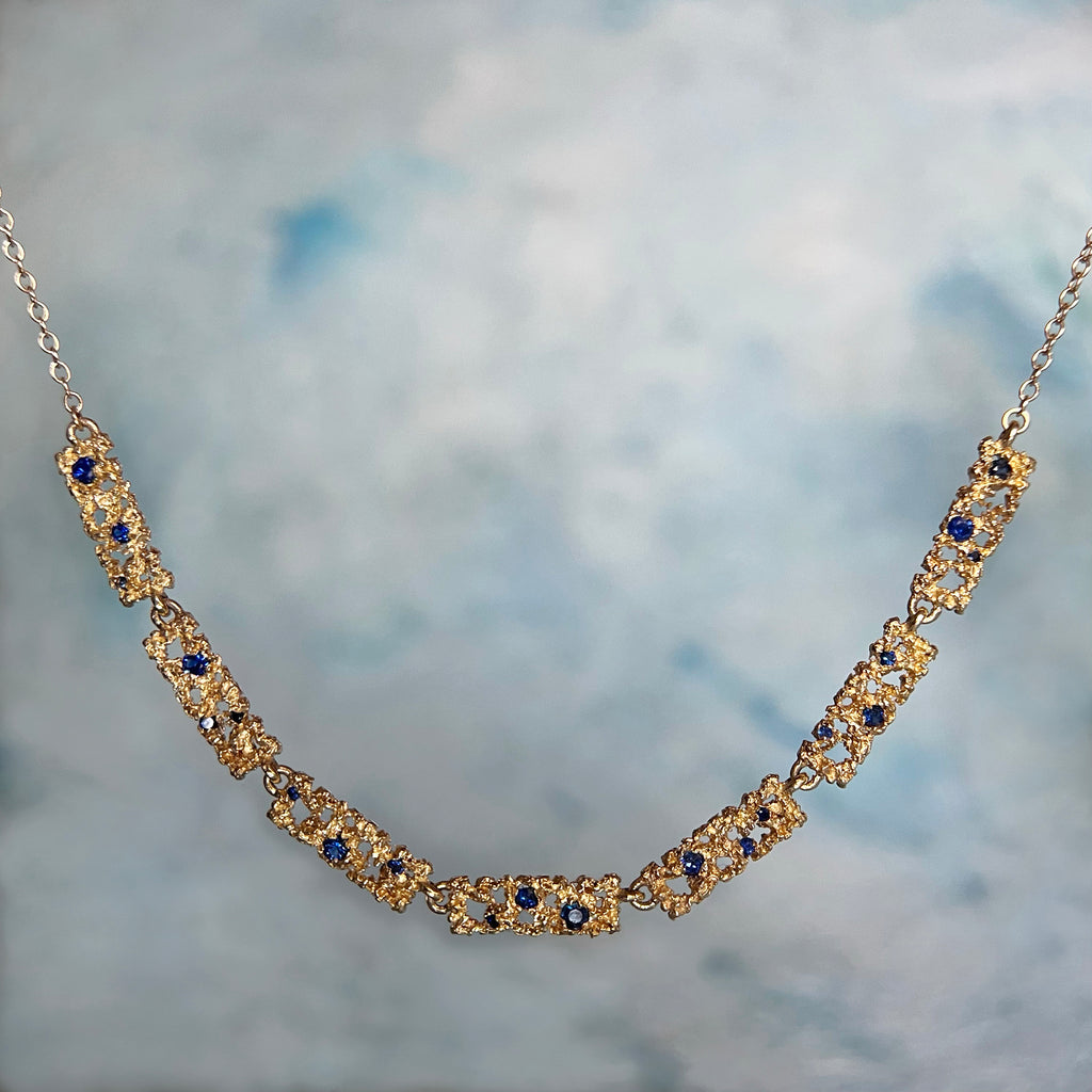 Seven textured and lacy rectangles form the bottom 25% of this necklace, with the rest made up of chain; shonw in 14k yellow with three blue sapphires scattered in each rectangle