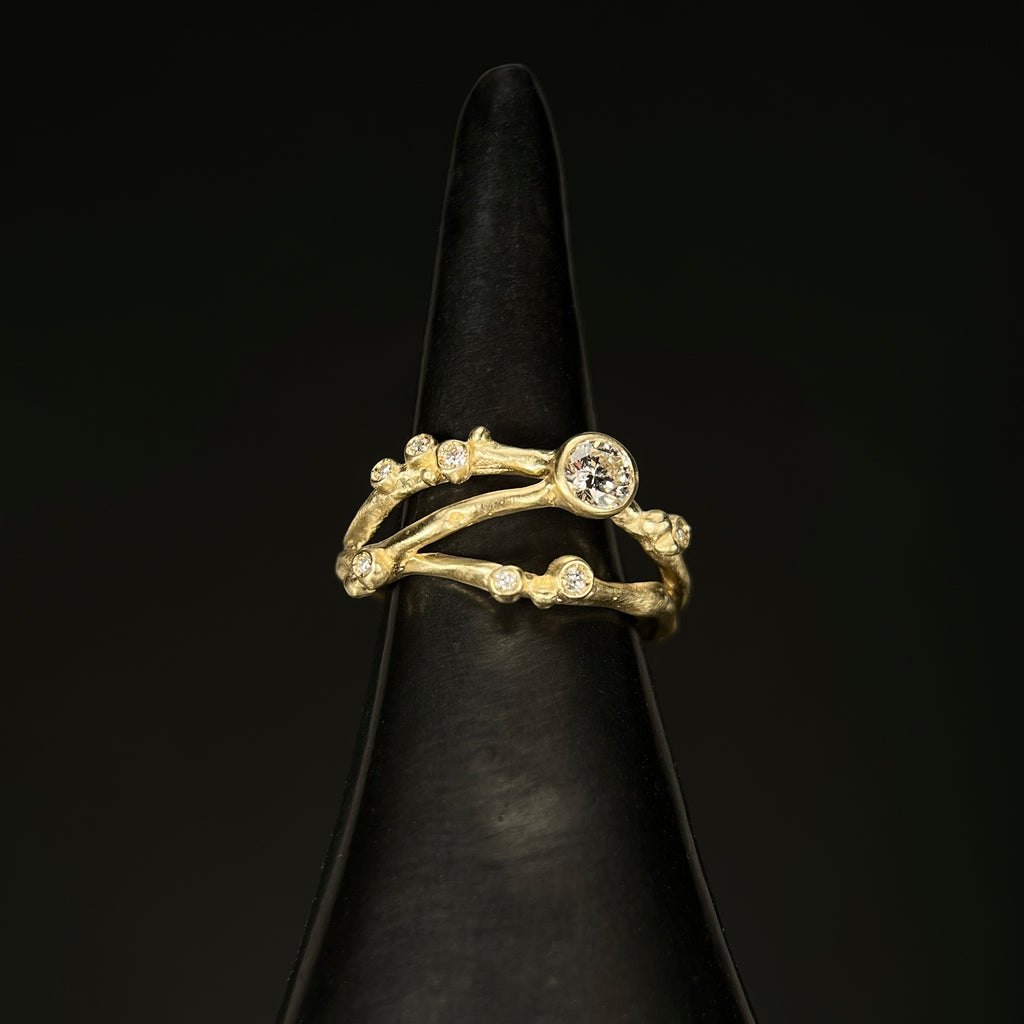 Organic, branch-like ring with three branches. Large off-center stone and seven tiny diamond buds. Shown in yellow gold on black background. 