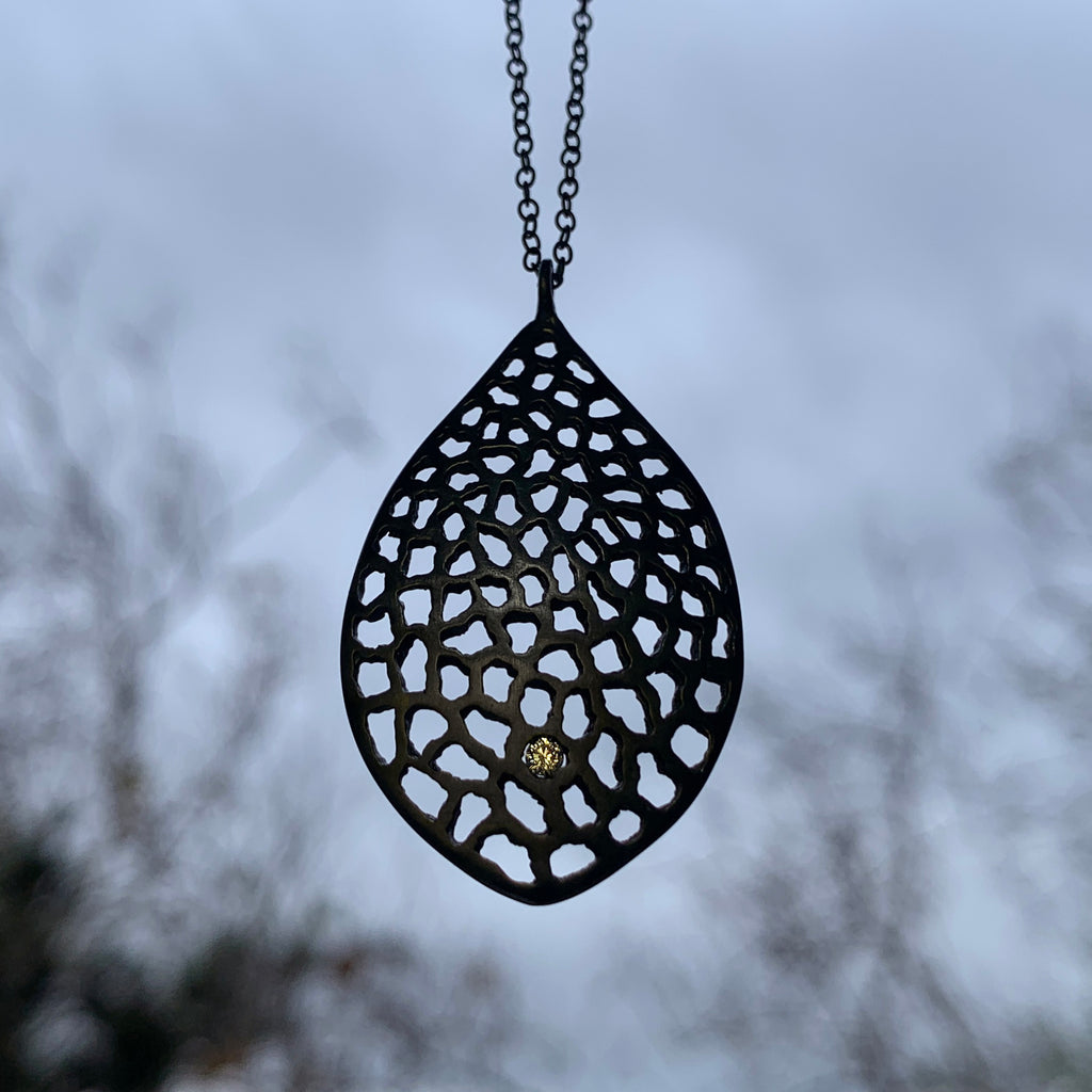 symmetrical, stylized leaf shaped pendant, filled with tiny, irregular cutouts and punctuated by a single diamond in the bottom quarter of the piece; shown in blackened silver; hanging in front of a window with grey skies and soft black branches
